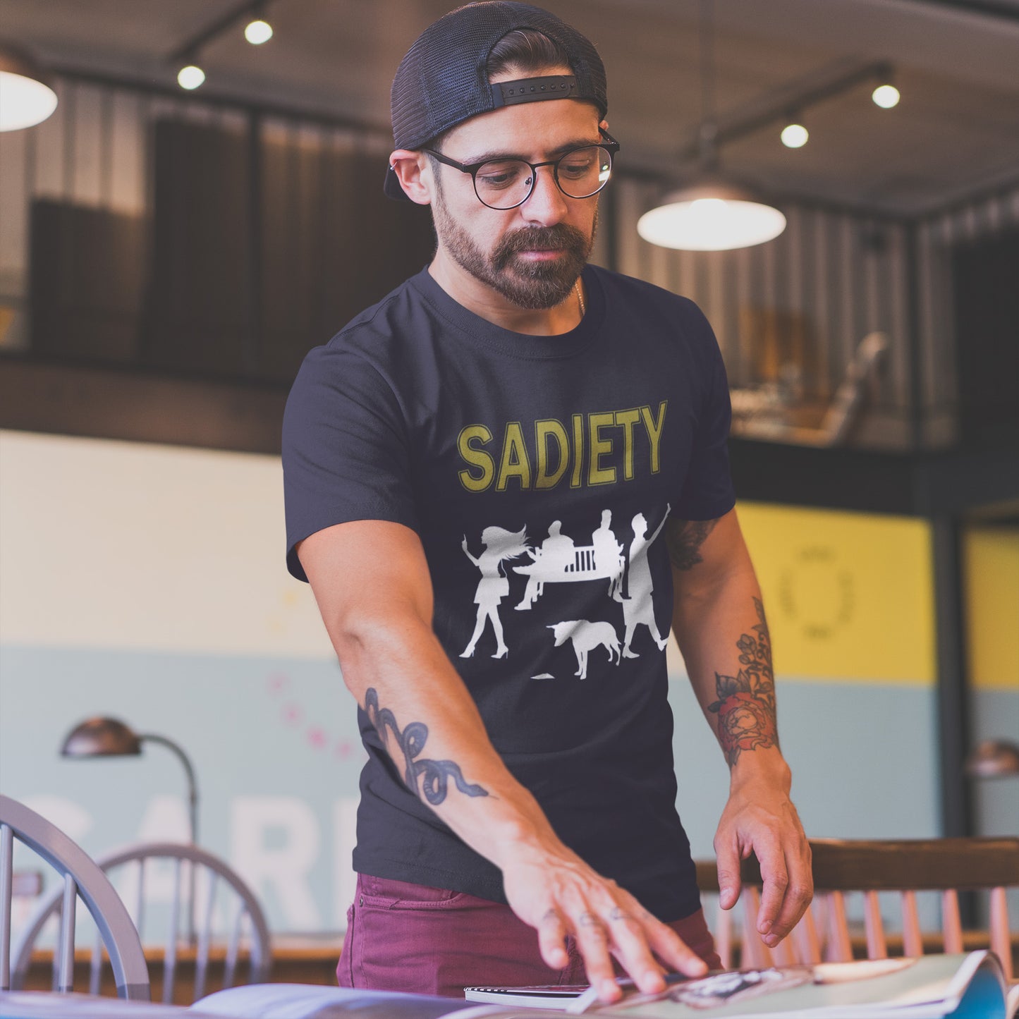 Man wearing a French navy t-shirt with 'SADIETY' design in a casual setting. A counterculture t-shirt design promoting social awareness about the effects of the mobile phone on society and sociability. The mobile phone has made people in close proximity seem miles apart. It's not a society, it's sadiety. Made from 100% organic ring-spun cotton, this subculture t-shirt is unisex, high-quality, super comfy, and best of all, eco-friendly.