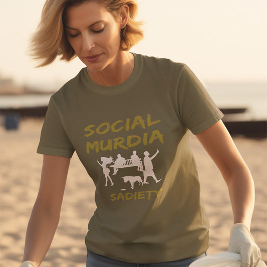 A woman on a beach wearing an khaki green t-shirt with a graphic print on the front. The print features the words 'SOCIAL MURDIA' in a textured gold font, a white silhouette of a group of people on smartphones and a dog, and the word 'SADIETY' in the same gold font below.
