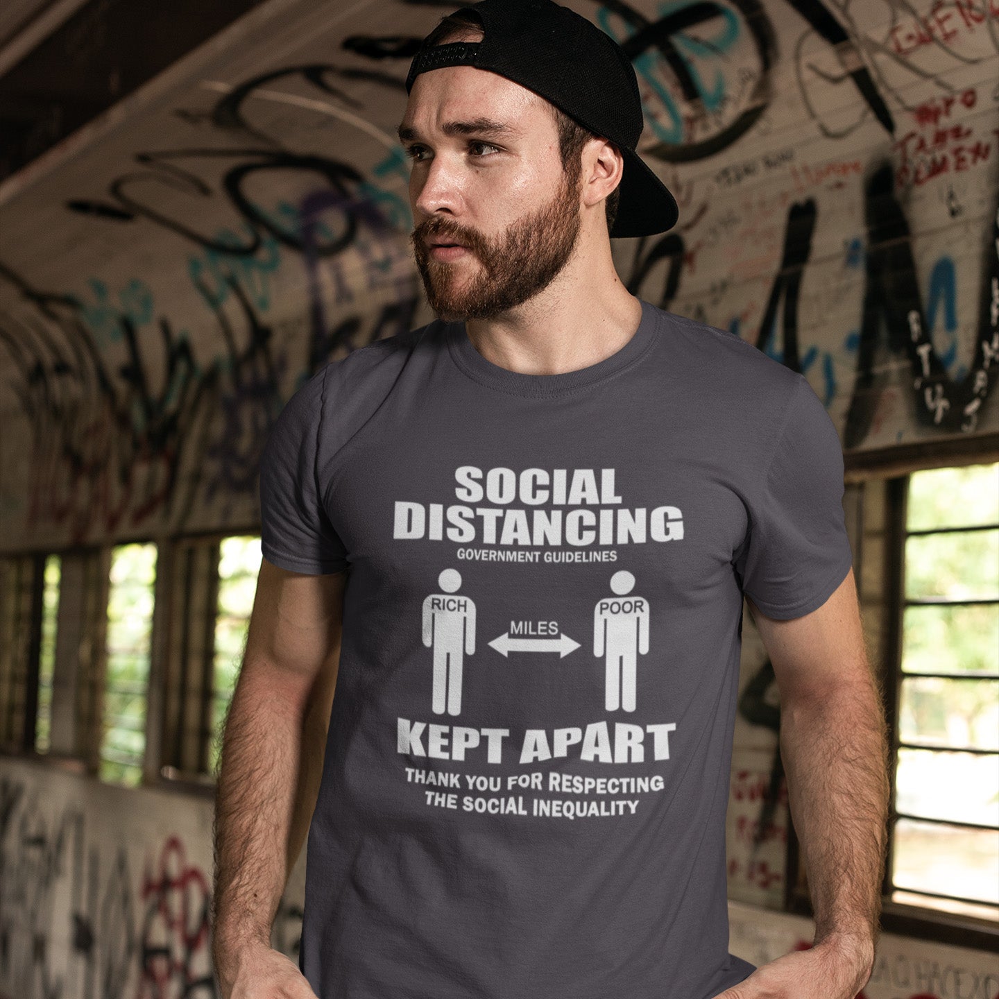 Man wearing a t-shirt with a social distancing graphic in an indoor setting with graffiti. A class war t-shirt design representing a much older form of social distancing in our society. Class inequality. This anti-establishment t-shirt design promote social awareness of the class divide our system creates. Made from 100% organic ring-spun cotton, this nonconformist t-shirt is unisex, high-quality, super comfy, and best of all, eco-friendly.