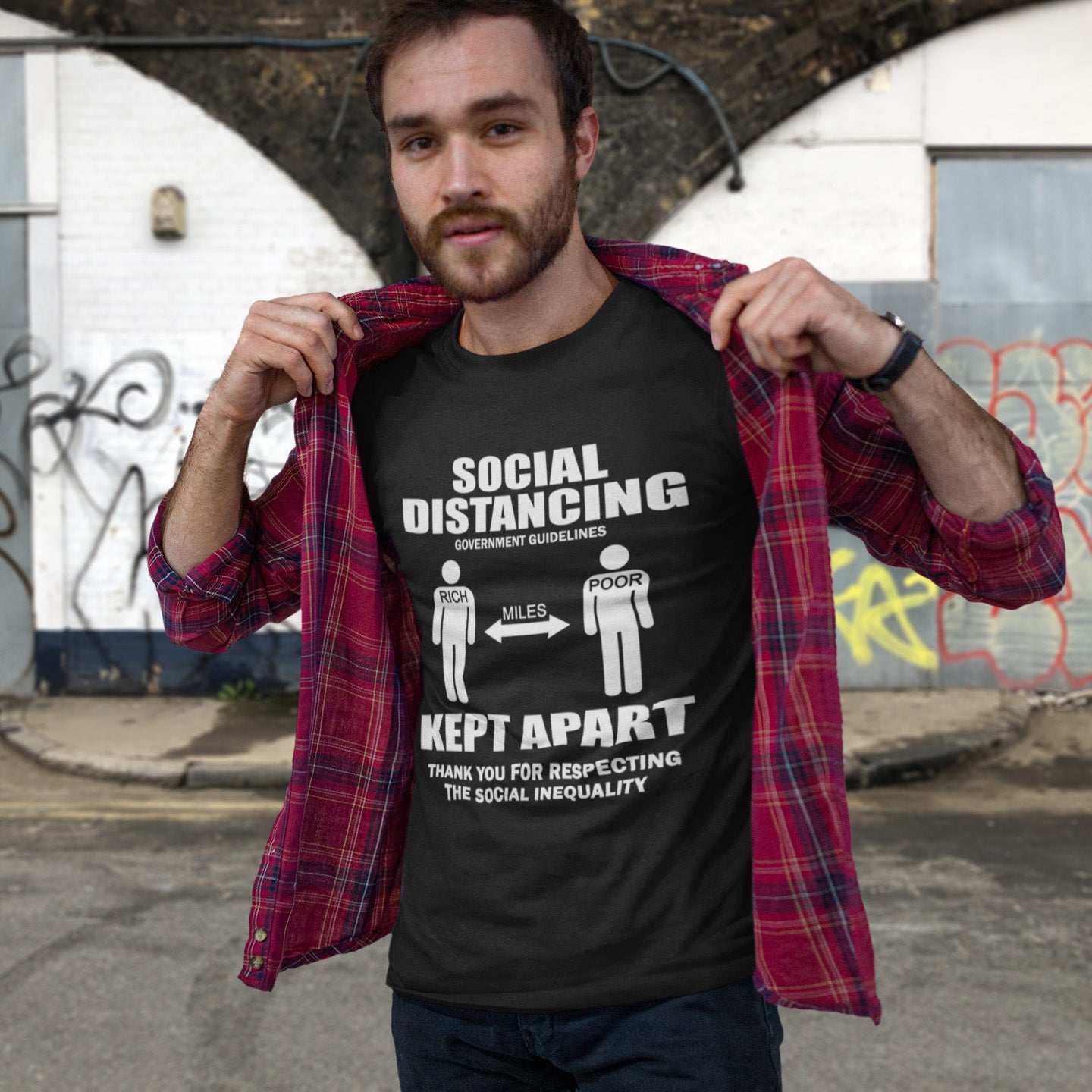 Man wearing a black t-shirt with social distancing graphic and text, standing against a graffiti-covered wall. A class war t-shirt design representing a much older form of social distancing in our society. Class inequality. This anti-establishment t-shirt design promote social awareness of the class divide our system creates. Made from 100% organic ring-spun cotton, this nonconformist t-shirt is unisex, high-quality, super comfy, and best of all, eco-friendly.
