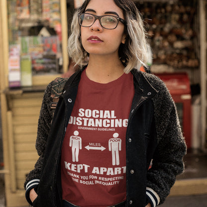 Person wearing a burgundy t-shirt with social distancing message in an indoor setting. A class war t-shirt design representing a much older form of social distancing in our society. Class inequality. This anti-establishment t-shirt design promote social awareness of the class divide our system creates. Made from 100% organic ring-spun cotton, this nonconformist t-shirt is unisex, high-quality, super comfy, and best of all, eco-friendly.