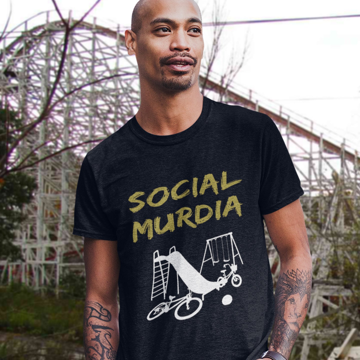 Man wearing a black t-shirt with 'SOCIAL MURDIA' graphic design in front of an amusement park. This counterculture t-shirt promoting social awareness of the effect of social media on our children. A unique design depicting how social media is probably murdering our society and leaving our playgrounds empty. Made from 100% organic ring-spun cotton, this anti-mobile phone t-shirt is unisex, high-quality, super comfy, and best of all, eco-friendly.