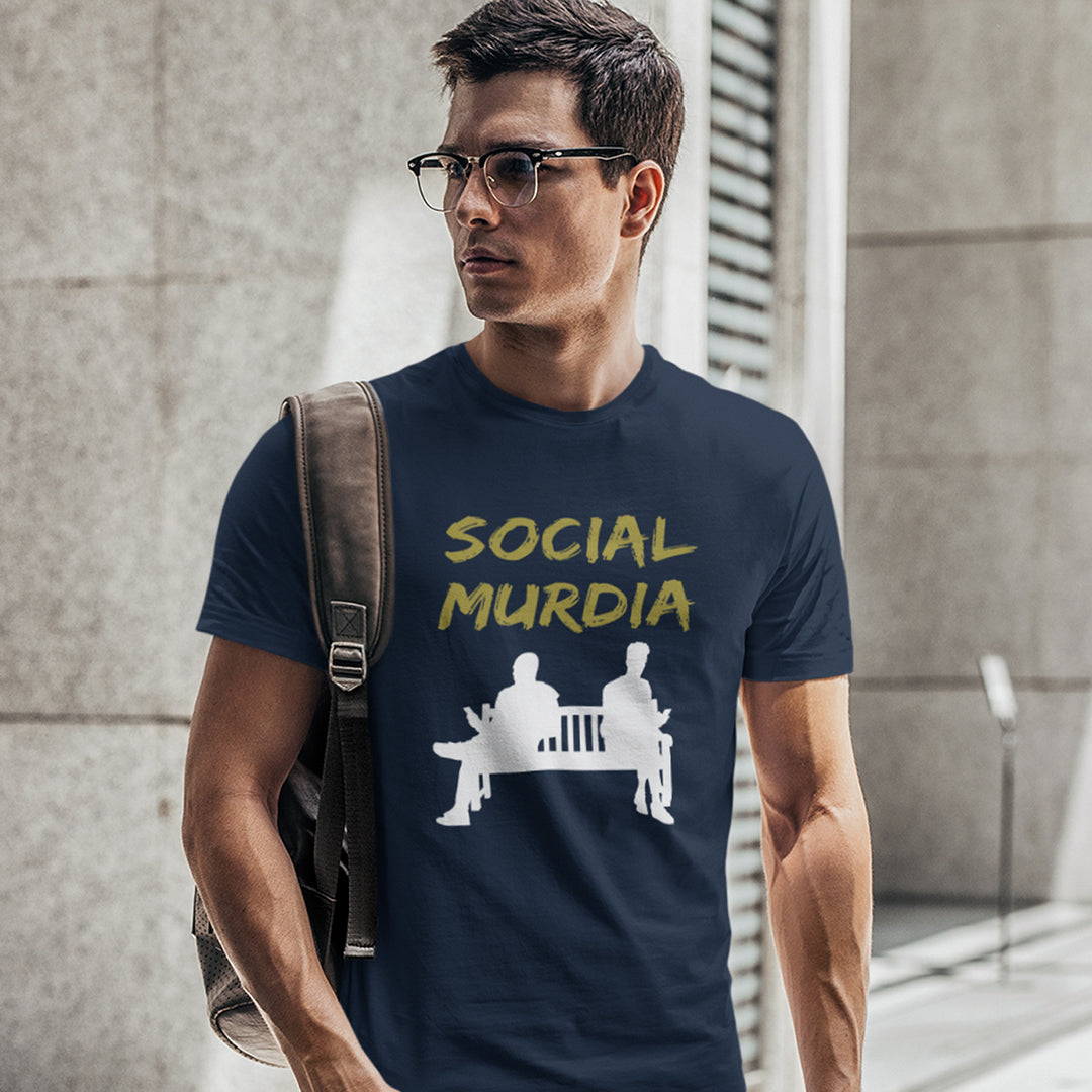 A man wearing a French navy blue graphic t-shirt with the words 'SOCIAL MURDIA' in yellow and a white graphic below, standing in front of a grey stone wall.