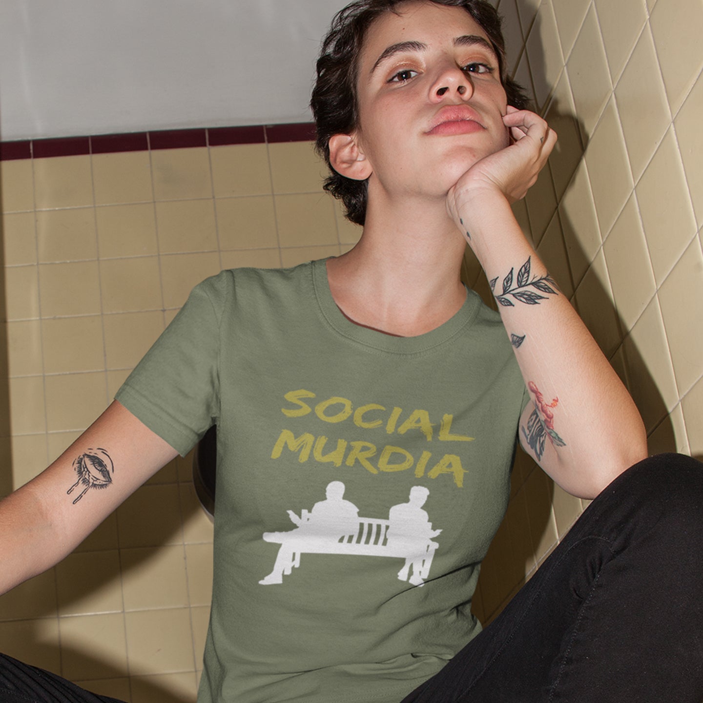 Person wearing a khaki t-shirt with 'SOCIAL MURDIA' text and graphic, sitting against a tiled wall. An anti-social media t-shirt arguing against the abuse of social media and mobile phones.  This anti-mobile phone design suggests society is actually being murdered by media on mobiles. Made from 100% organic ring-spun cotton, this anti-social media t-shirt is unisex, high-quality, super comfy, and best of all, eco-friendly.