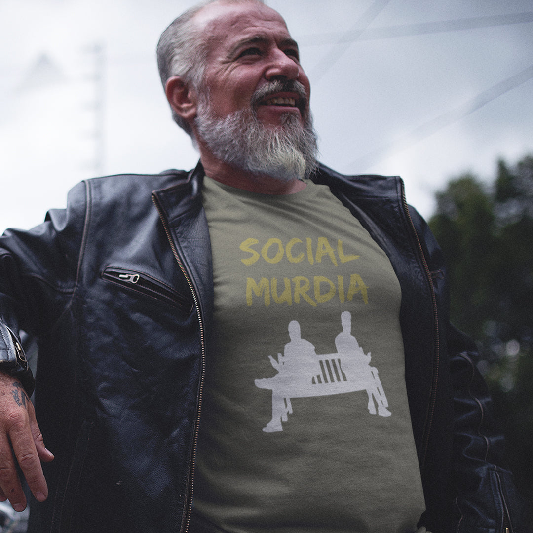 A smiling older man with a grey beard wearing a black leather jacket over an khaki green graphic t-shirt that reads 'SOCIAL MURDIA' with a white bench graphic, photographed outdoors against a blurred sky and trees.