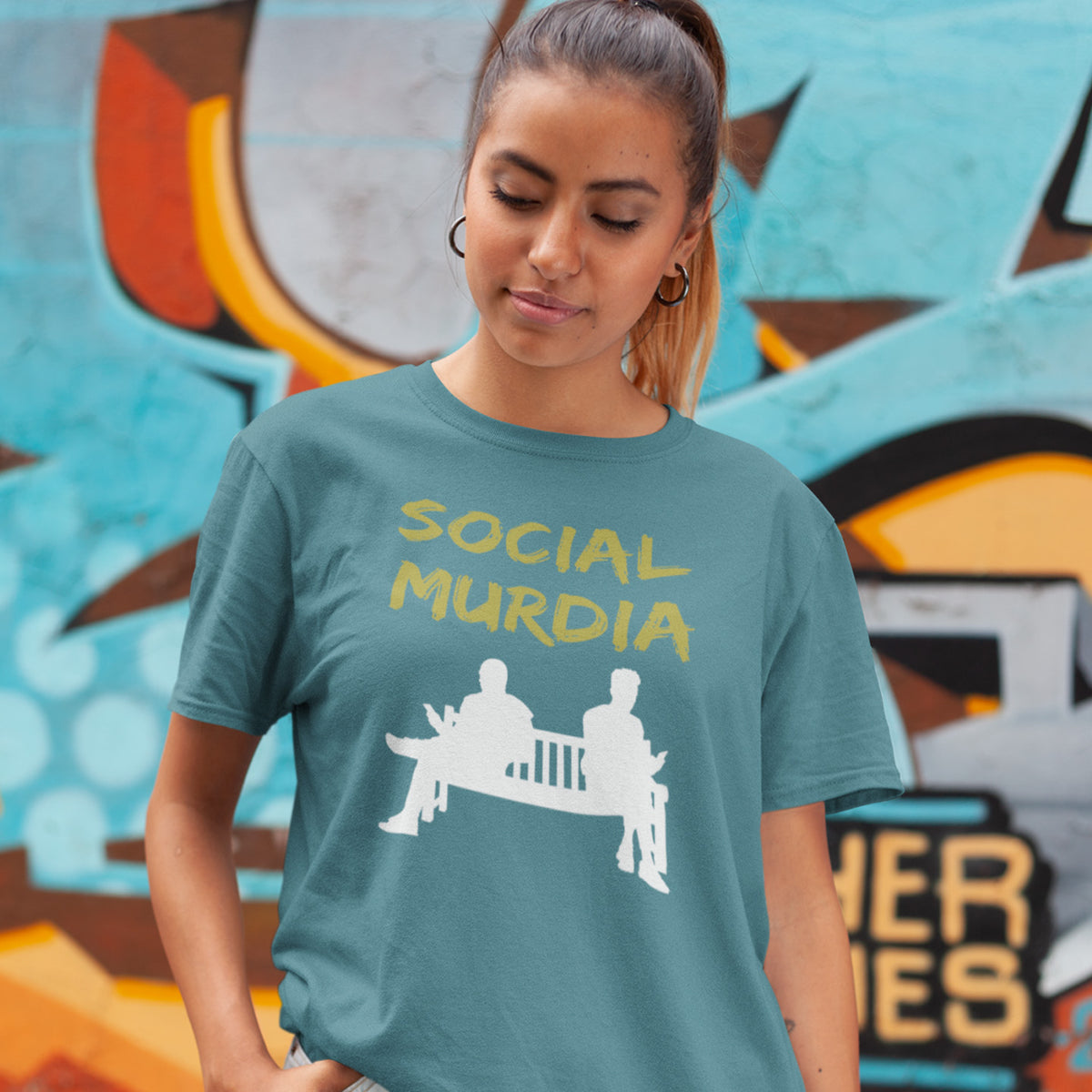 A woman wearing a stargazer blue t-shirt with a yellow and white graphic that reads 'SOCIAL MURDIA' and shows two silhouettes sitting on a bench. She is standing in front of a colourful graffiti wall.