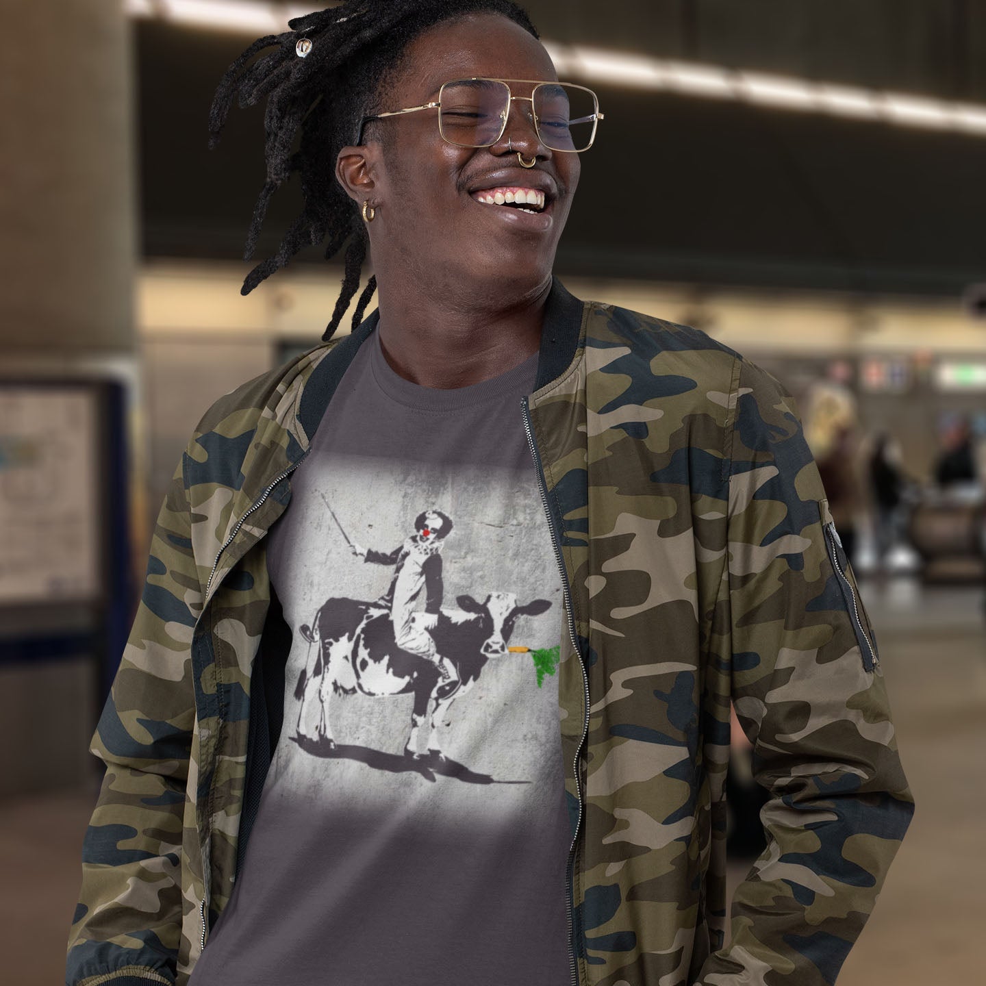 Person wearing a camouflage jacket over a graphic t-shirt with a cow and rider design. A stencil graffiti t shirt. A metaphor for a system of motivation. The 'stick or carrot' argument portrayed in a street art stencil design. Made from 100% organic ring-spun cotton, this urban art t shirt is unisex, high-quality, super comfy, and best of all, eco-friendly.