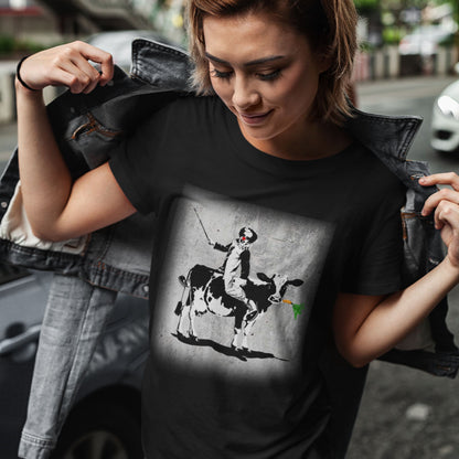 Person wearing a black t-shirt with a graphic design of a person riding a cow, standing on a street. A stencil graffiti t shirt. A metaphor for a system of motivation. The 'stick or carrot' argument portrayed in a street art stencil design. Made from 100% organic ring-spun cotton, this urban art t shirt is unisex, high-quality, super comfy, and best of all, eco-friendly.