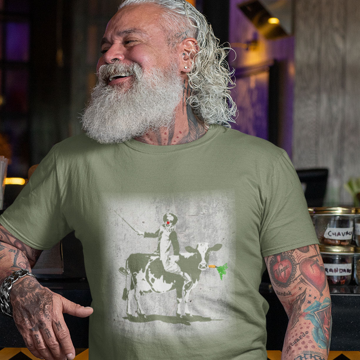 Man wearing a khaki t-shirt with a graphic design of a person on a cow, sitting in a bar. A stencil graffiti t shirt. A metaphor for a system of motivation. The 'stick or carrot' argument portrayed in a street art stencil design. Made from 100% organic ring-spun cotton, this urban art t shirt is unisex, high-quality, super comfy, and best of all, eco-friendly.