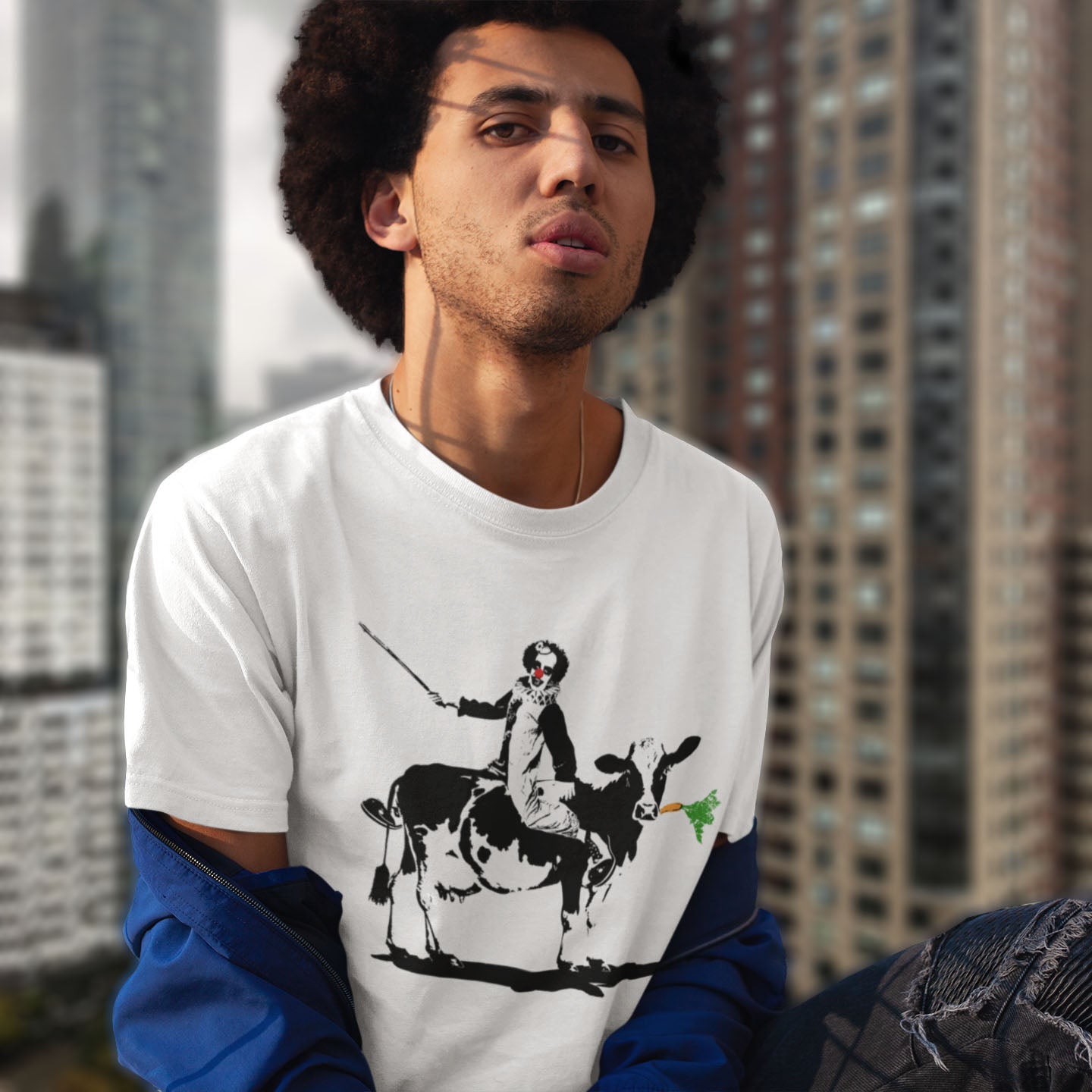 Person wearing a white t-shirt with a graphic design of a person riding a cow, standing in an urban setting. A stencil graffiti t shirt. A metaphor for a system of motivation. The 'stick or carrot' argument portrayed in a street art stencil design. Made from 100% organic ring-spun cotton, this urban art t shirt is unisex, high-quality, super comfy, and best of all, eco-friendly.
