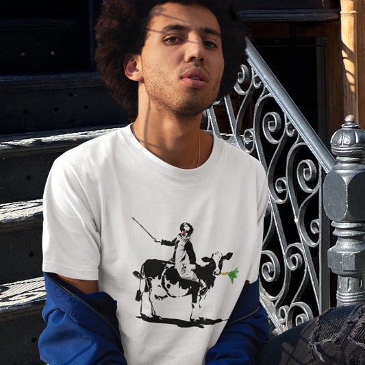 Person wearing a white t-shirt with a graphic design of a person riding a cow, standing in an urban setting. A stencil graffiti t shirt. A metaphor for a system of motivation. The 'stick or carrot' argument portrayed in a street art stencil design. Made from 100% organic ring-spun cotton, this urban art t shirt is unisex, high-quality, super comfy, and best of all, eco-friendly.