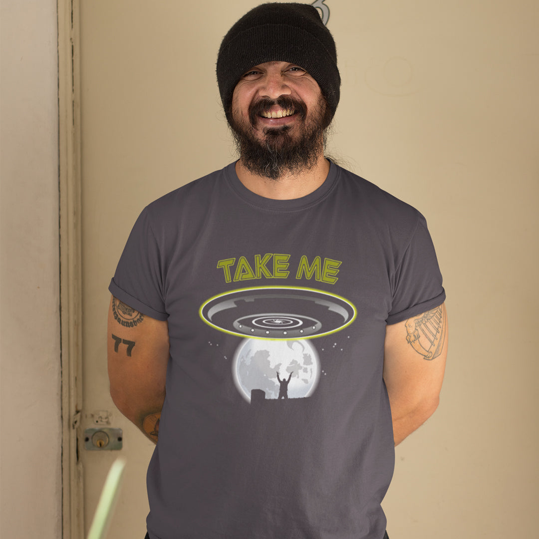A man wearing an anthracite grey t-shirt with a graphic of a UFO and the text 'TAKE ME' under it, standing against a beige wall.