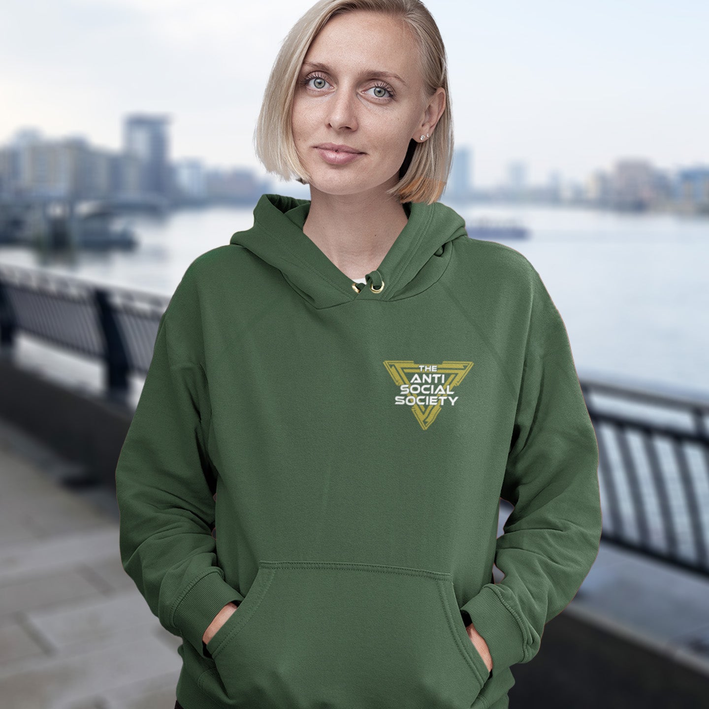 Person wearing a bottle green hoodie with a logo, standing by a waterfront with a cityscape in the background. This anti-social hoodie design gives you an oxymoron to ponder, the more and more you don't want to be a member, the more you become one. A satirical counterculture dig at modern society. Comfortable and practical. It's extra soft and has a convenient front pouch pocket. Made from organic cotton and recycled polyester, this social awareness hoodie is a great eco-friendly choice.