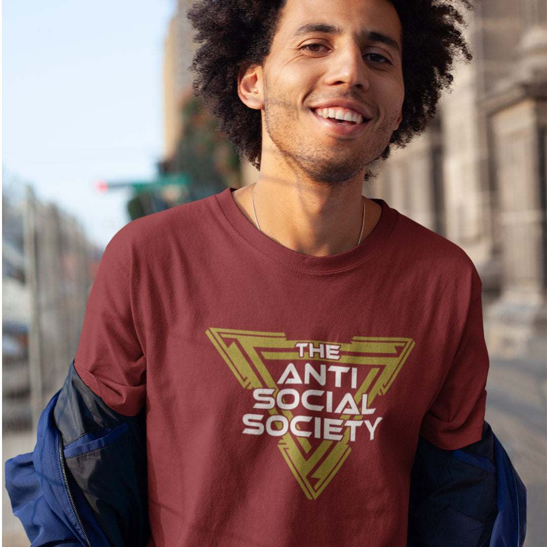 A smiling man wearing a burgundy crewneck t-shirt with a gold and white graphic that reads 'THE ANTI SOCIAL SOCIETY' inside a geometric emblem. He has a blue jacket draped off his shoulders and is photographed outdoors with a blurred city street background.