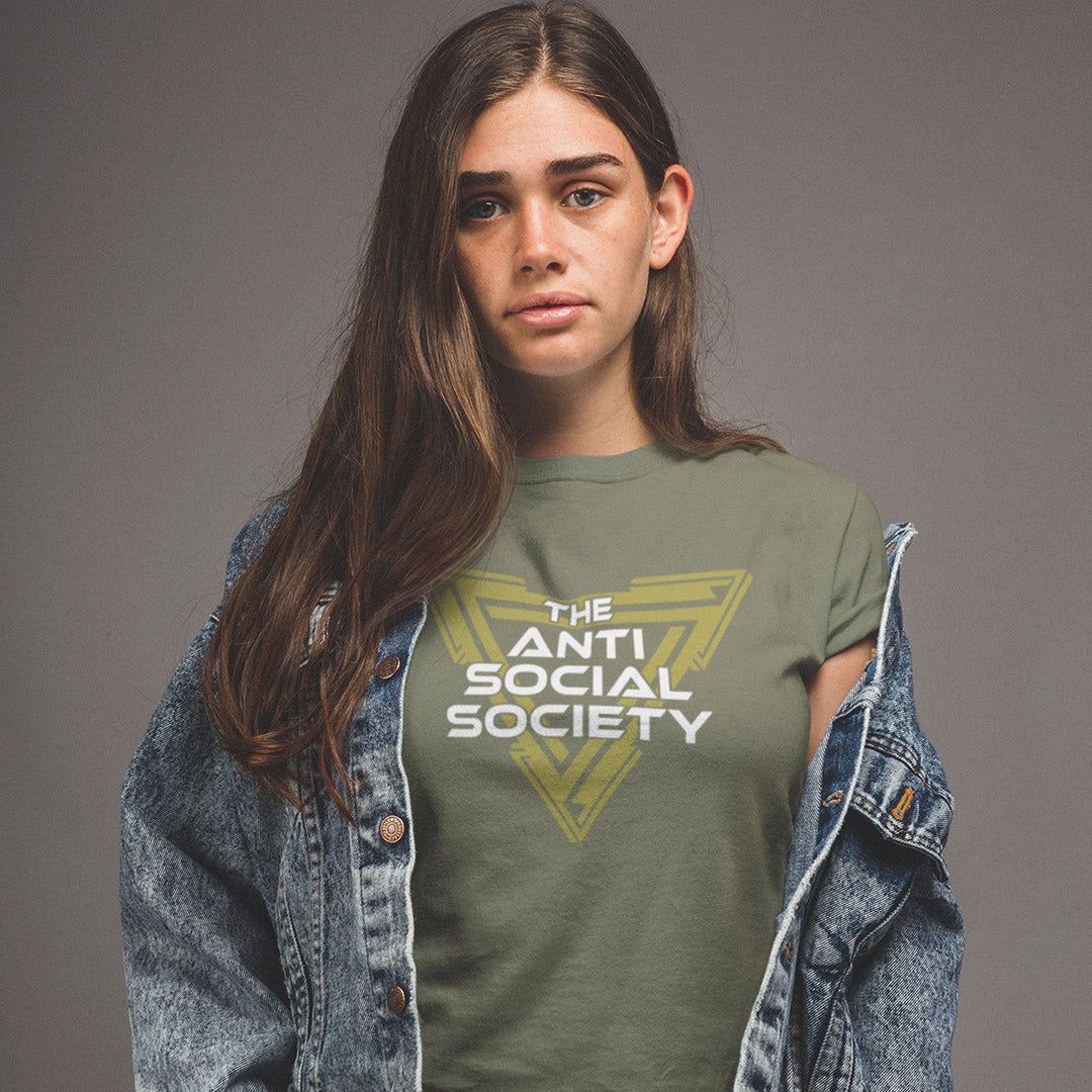 A woman wearing a khaki green graphic t-shirt with a yellow and white 'The Anti Social Society' logo, styled with an open blue denim jacket against a solid gey studio background.