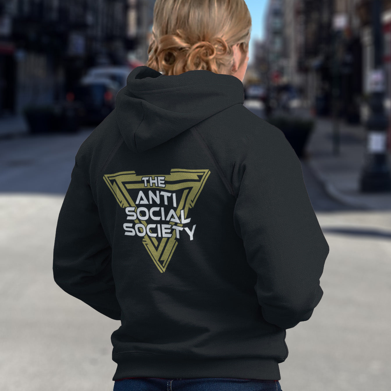 Person wearing a black hoodie with 'The Anti Social Society' logo on the back, standing on a city street. This anti-social clothing design gives you an oxymoron to ponder, the more and more you don't want to be a member, the more you become one. A satirical counterculture dig at modern society. Comfortable and practical. It's extra soft and has a convenient front pouch pocket. Made from organic cotton and recycled polyester, this social awareness hoodie is a great eco-friendly choice.