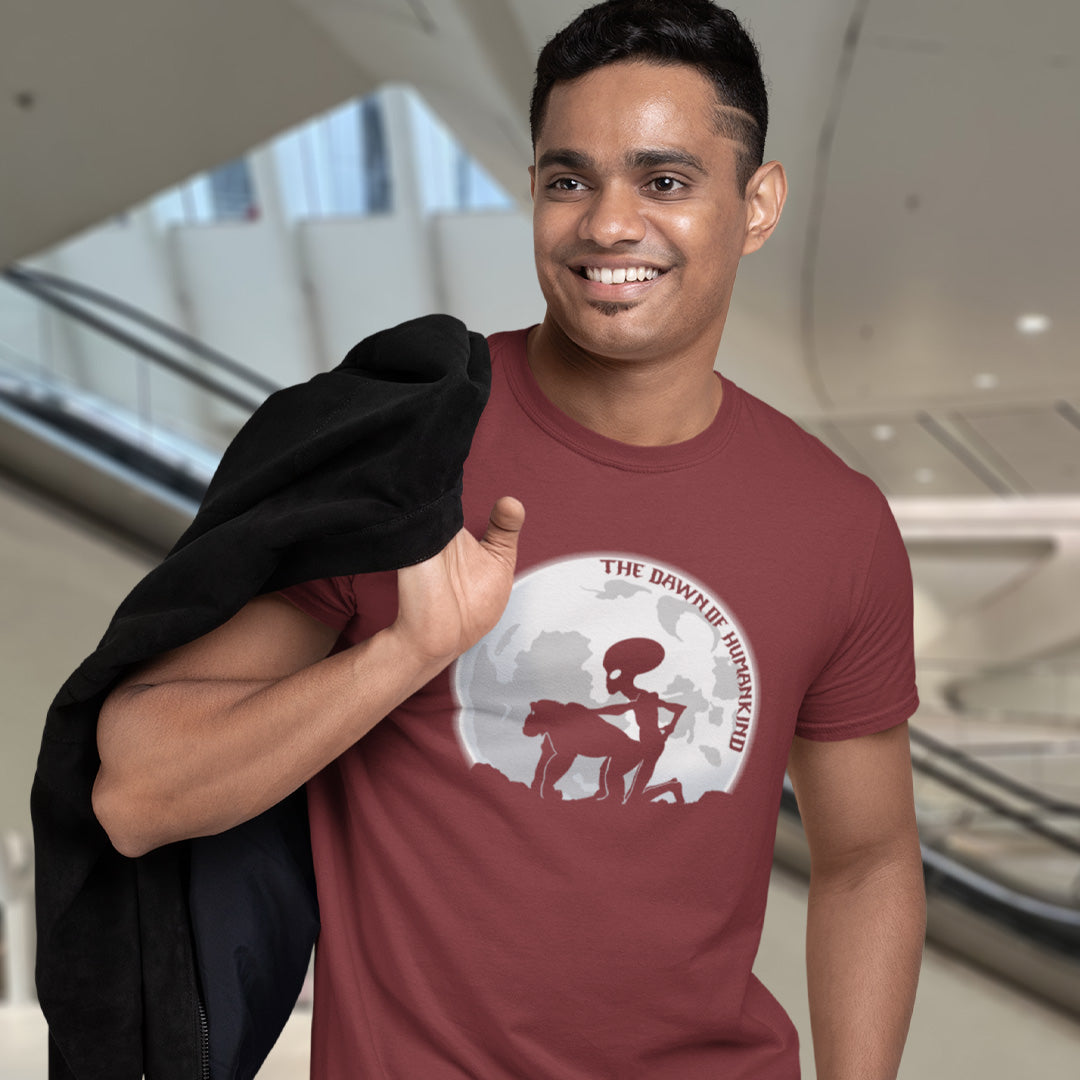 A man in a shopping centre, smiling and wearing a burgundy, short sleeved, crew neck t-shirt with the silhouette of an alien and a monkey in front of the moon with the text "THE DAWN OF HUMANKIND".