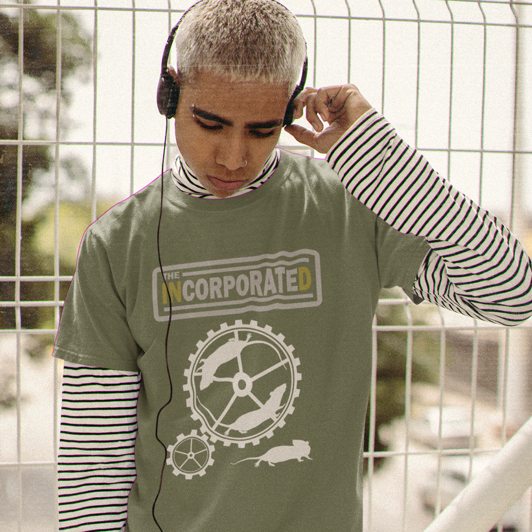 Person wearing a khaki t-shirt with a logo and text, standing in front of a wire fence. This non conformist design questions the effects of a corporate run society in the form of a spoof rock band t shirt. Have we just created a corporate cult to follow ? A satirical and alternative view of the modern rat race. Made from 100% organic ring-spun cotton, this counter culture t shirt is unisex, high-quality, super comfy, and best of all, eco-friendly.