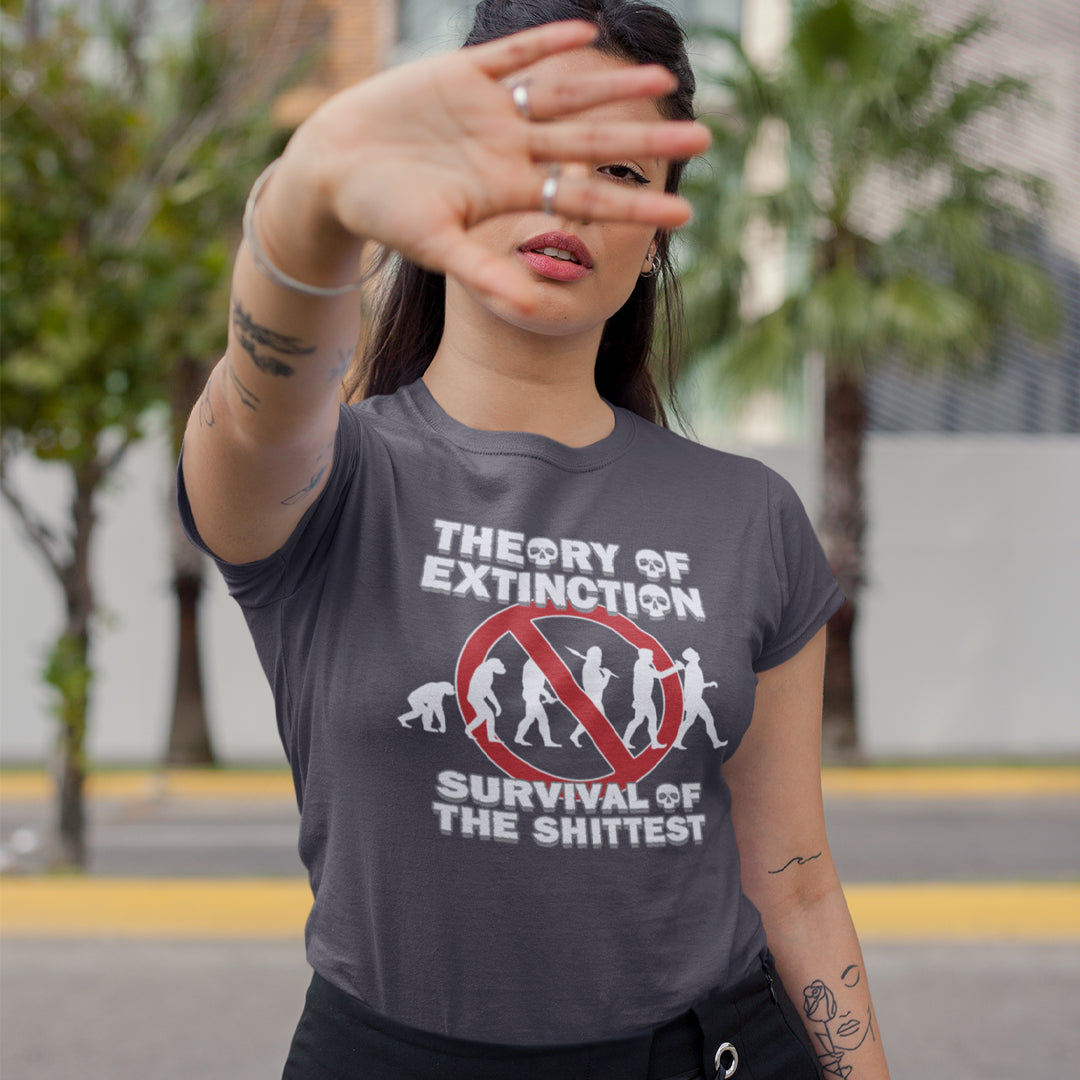 Woman outside with her hand up, wearing an anthracite grey t-shit with a graphic on the front. The graphic features the text 'THEORY OF EXTINCTION' and 'SURVIVAL OF THE SHITTEST' in white, surrounding a red prohibition sign over white silhouettes of human evolution.