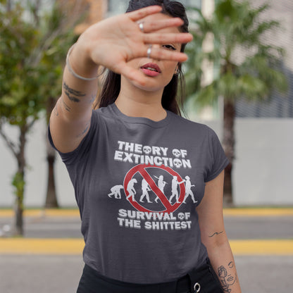 Woman outside with her hand up, wearing an anthracite grey t-shit with a graphic on the front. The graphic features the text 'THEORY OF EXTINCTION' and 'SURVIVAL OF THE SHITTEST' in white, surrounding a red prohibition sign over white silhouettes of human evolution.