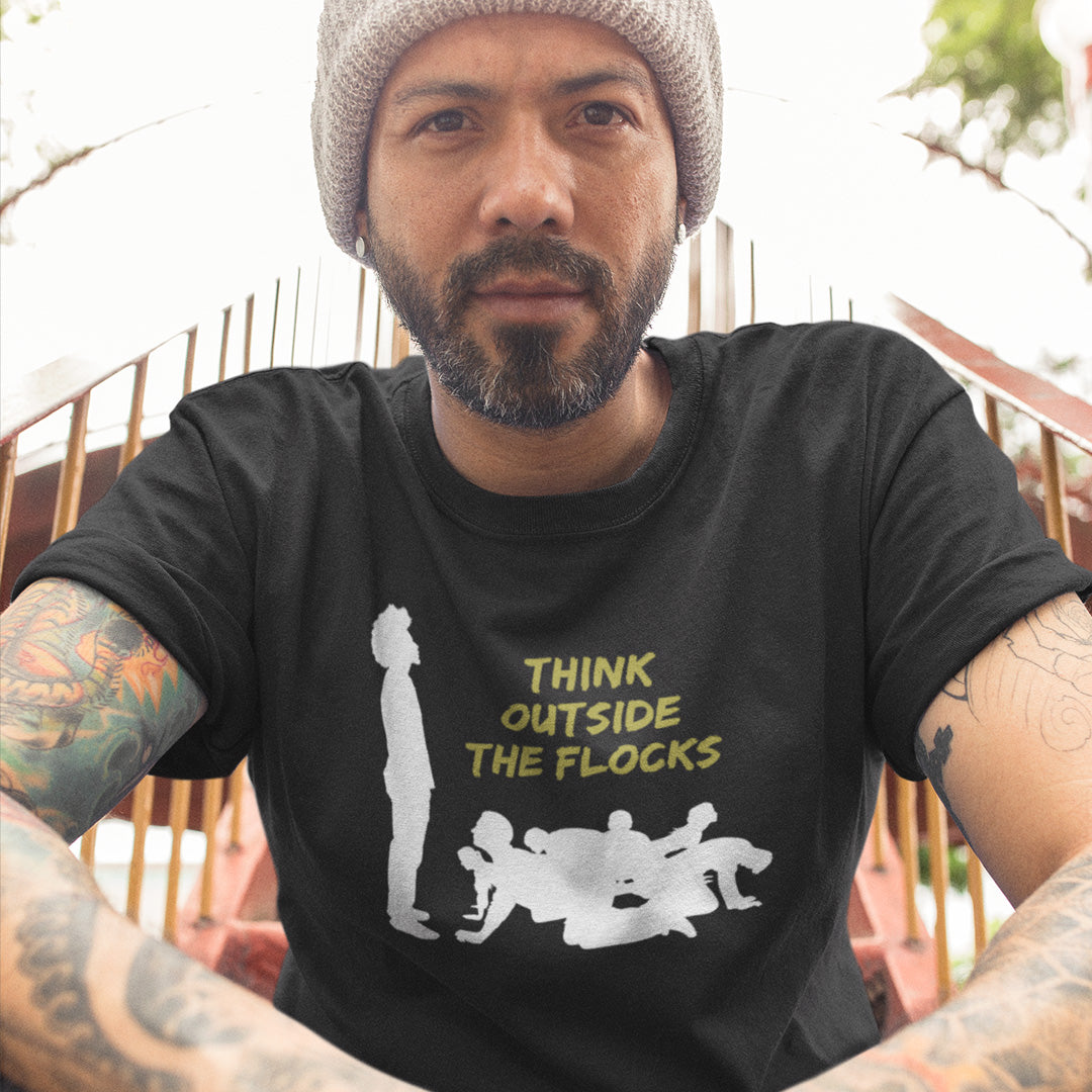 A man wearing a black graphic t-shirt with a white silhouette of sheep and yellow text reading 'THINK OUTSIDE THE FLOCKS', paired with a gray knit beanie. The photo is taken from a low angle, showing tattooed arms in the foreground and a fence in the background.