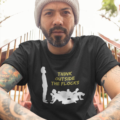 A man wearing a black graphic t-shirt with a white silhouette of sheep and yellow text reading 'THINK OUTSIDE THE FLOCKS', paired with a gray knit beanie. The photo is taken from a low angle, showing tattooed arms in the foreground and a fence in the background.