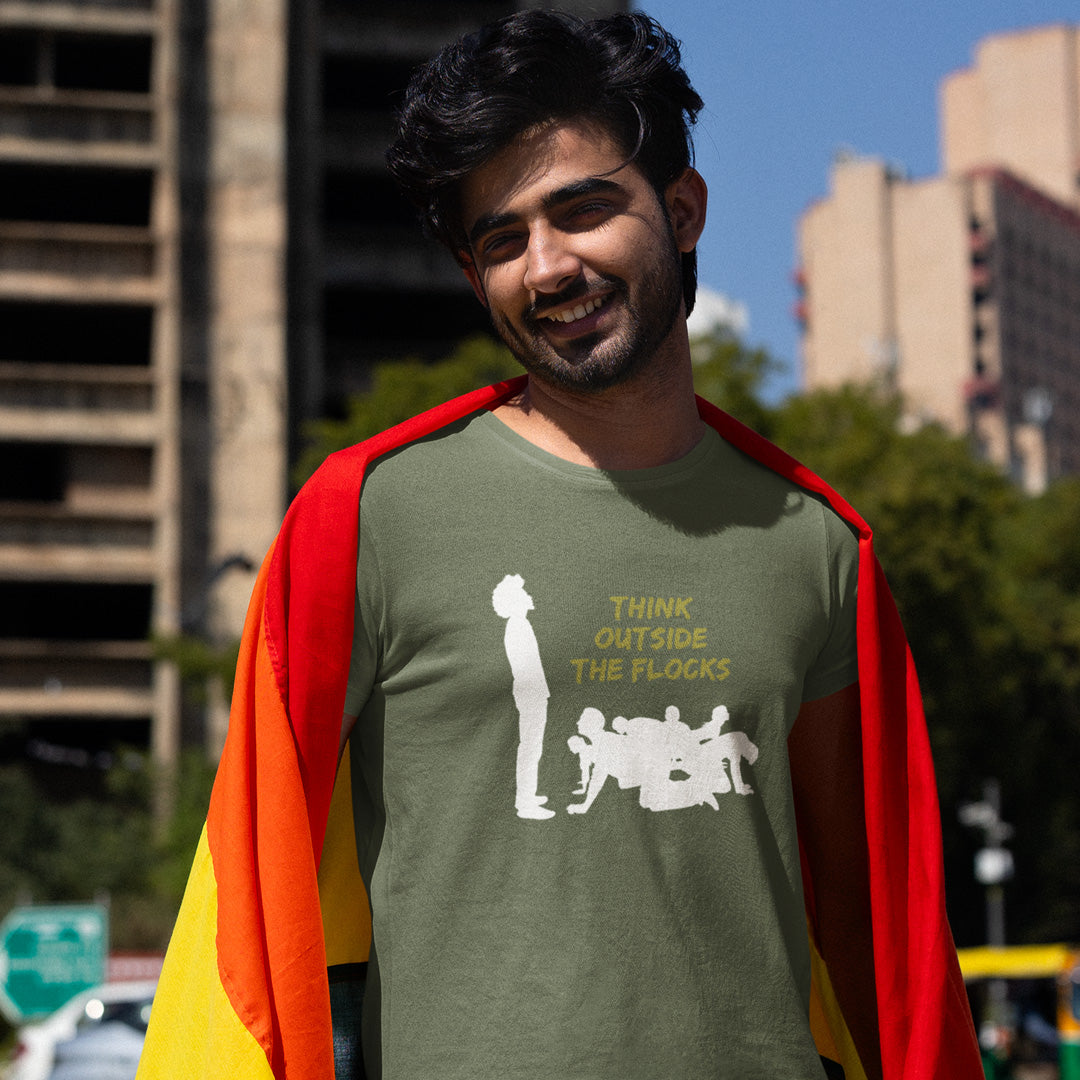 A man outdoors wearing an olive green graphic T-shirt with a white silhouette and yellow text reading 'THINK OUTSIDE THE FLOCKS,' and draped over his shoulders are red, orange, and yellow flag-like panels. Buildings and trees are blurred in the background.