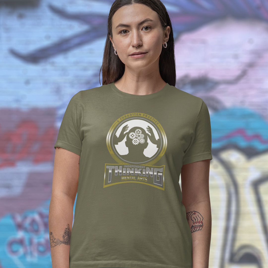 A woman wearing an olive green t-shirt with a gold and white 'Thinking Mentally Arts' logo on the chest, standing against a blurred graffiti wall.