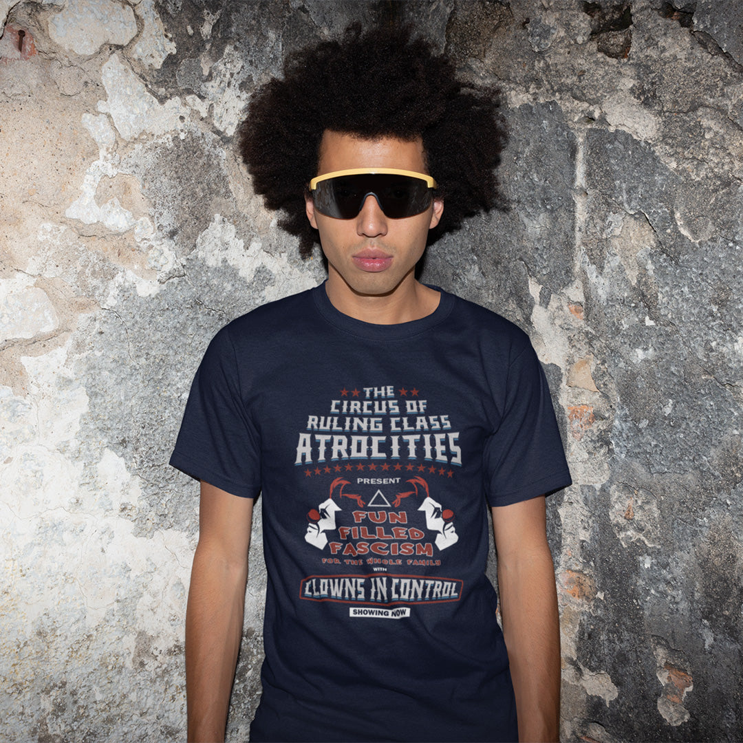A man in sunglasses, standing against a rough wall, wearing a French navy blue short sleeved crew neck t-shirt with a spoof circus poster design named "The Circus of Ruling Class Atrocities" and "Clowns in Control" text.