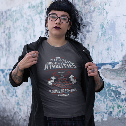 A tattooed woman in glasses, standing against an unfinished concrete wall, holding open her jacket to reveal an anthracite grey short sleeved crew neck t-shirt with a spoof circus poster design named "The Circus of Ruling Class Atrocities" and "Clowns in Control" text.