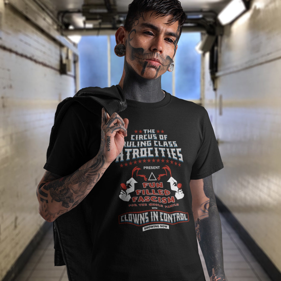 A heavily tattooed man standing in a public corridor with his jacket over his shoulder, wearing a black short sleeved crew neck t-shirt with a spoof circus poster design named "The Circus of Ruling Class Atrocities" and "Clowns in Control" text.