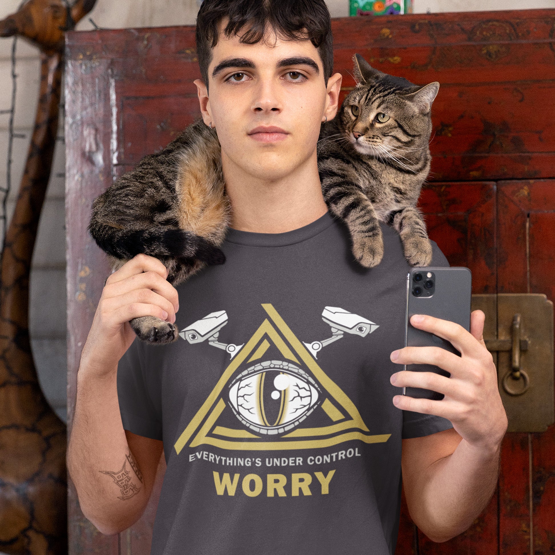 Man wearing a t-shirt with a cat on his shoulders and holding a phone, standing in front of a wooden door.
