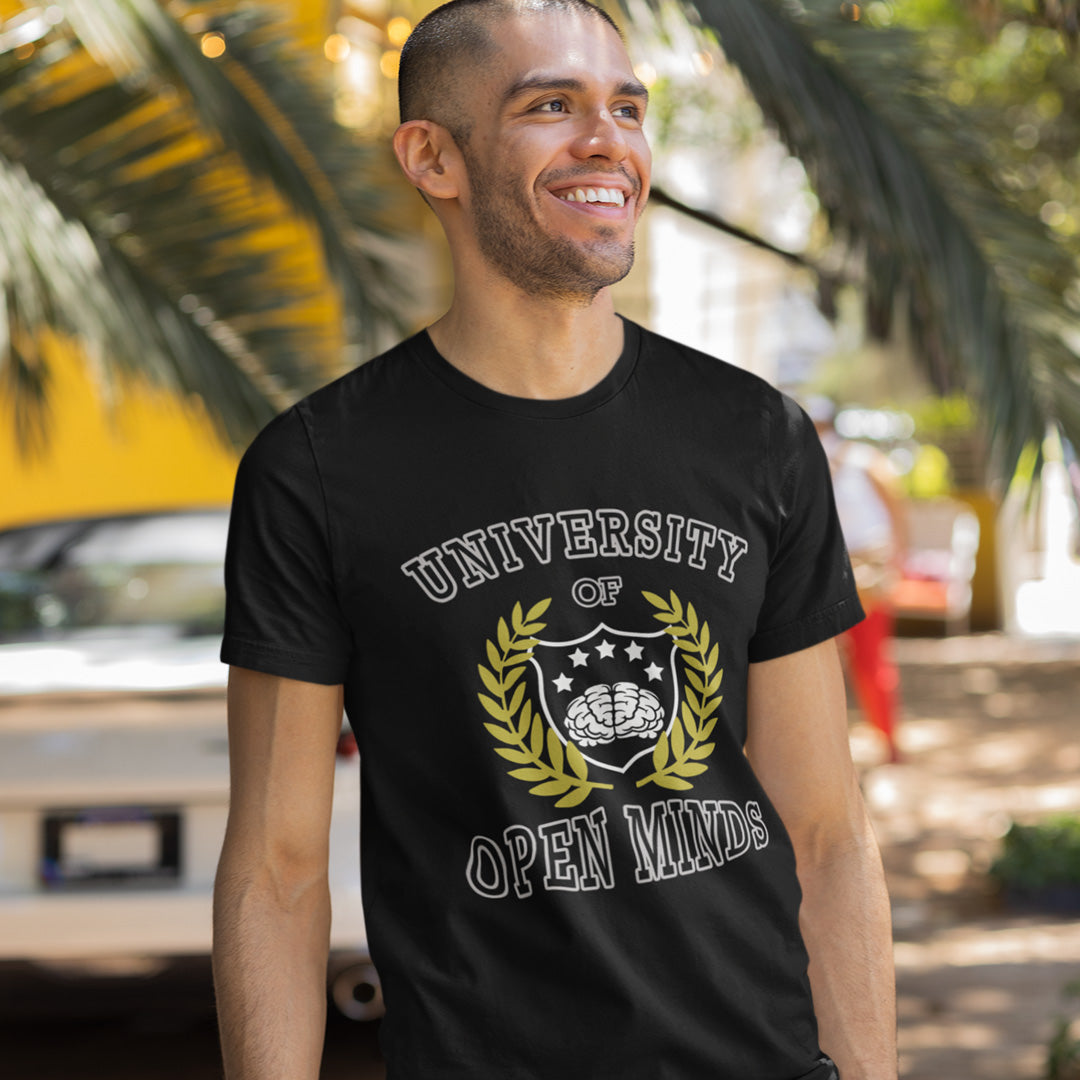 A man wearing a black graphic t-shirt with the text 'UNIVERSITY OF OPEN MINDS' and a laurel wreath design, set against a blurred outdoor background with palm trees.