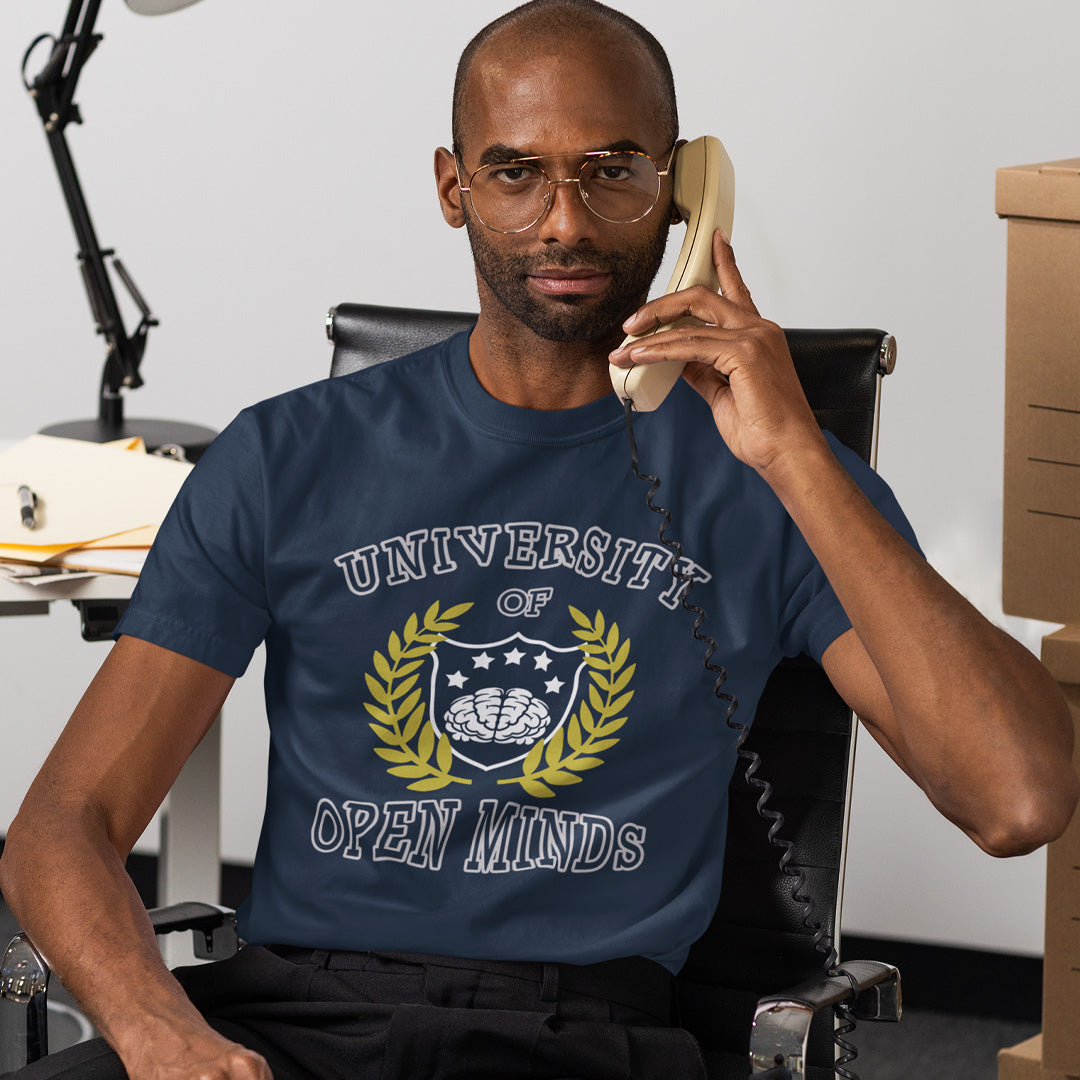 A man wearing a navy blue graphic t-shirt with a 'University of Open Minds' emblem, sitting in an office chair and holding a vintage telephone receiver.