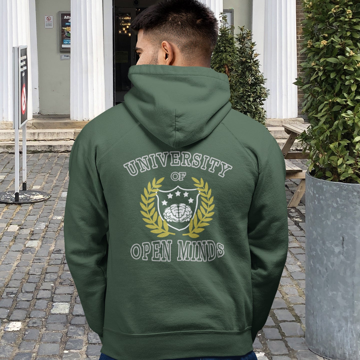 Person wearing a green hoodie with 'University of Open Minds' design outdoors. A graphic hoodie design for the liberal minded. If you think outside the box then this university is for you. Only practical lessons and no student loan required. Comfortable and practical. It's extra soft and has a convenient front pouch pocket. Made from organic cotton and recycled polyester, this graphic hoodie is a great eco-friendly choice.