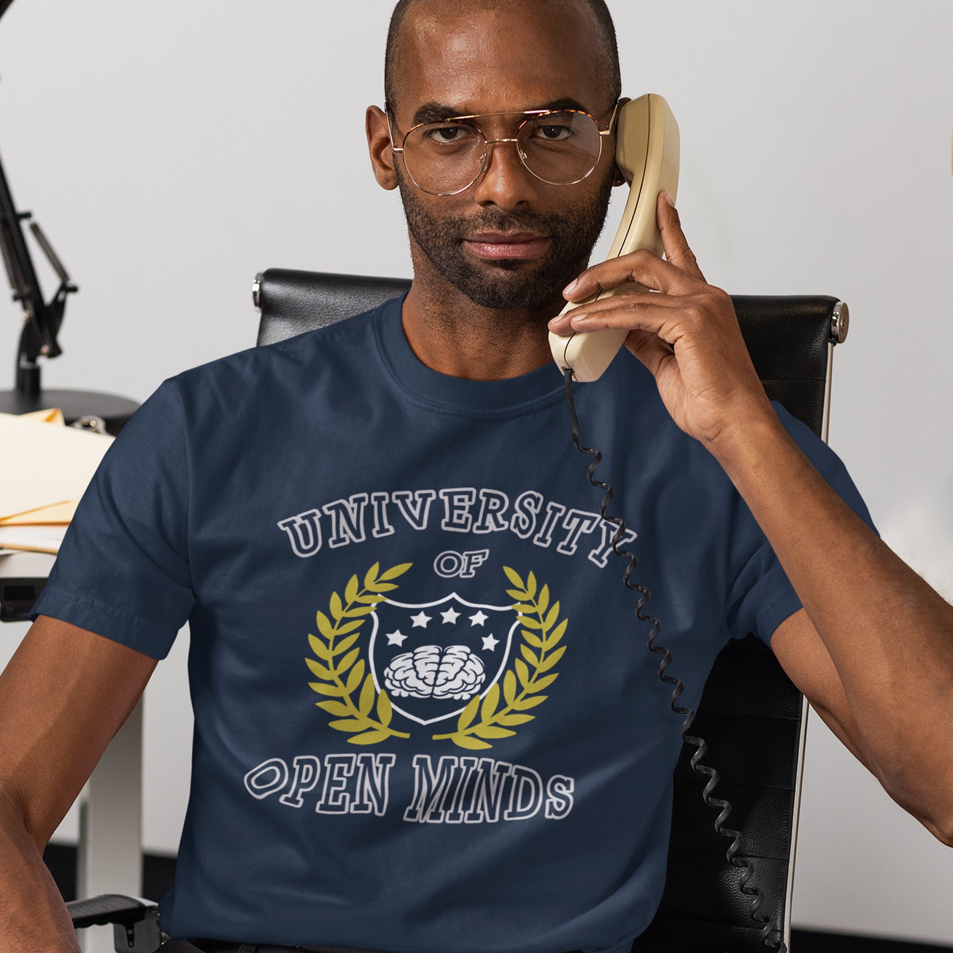 Man wearing a navy blue t-shirt with 'University of Open Minds' design, sitting in an office chair. A cool graphic design for the liberal minded. If you think outside the box then this university is for you. Only practical lessons and no student loan required. Made from 100% organic ring-spun cotton, this social awareness t-shirt is unisex, high-quality, super comfy, and best of all, eco-friendly.