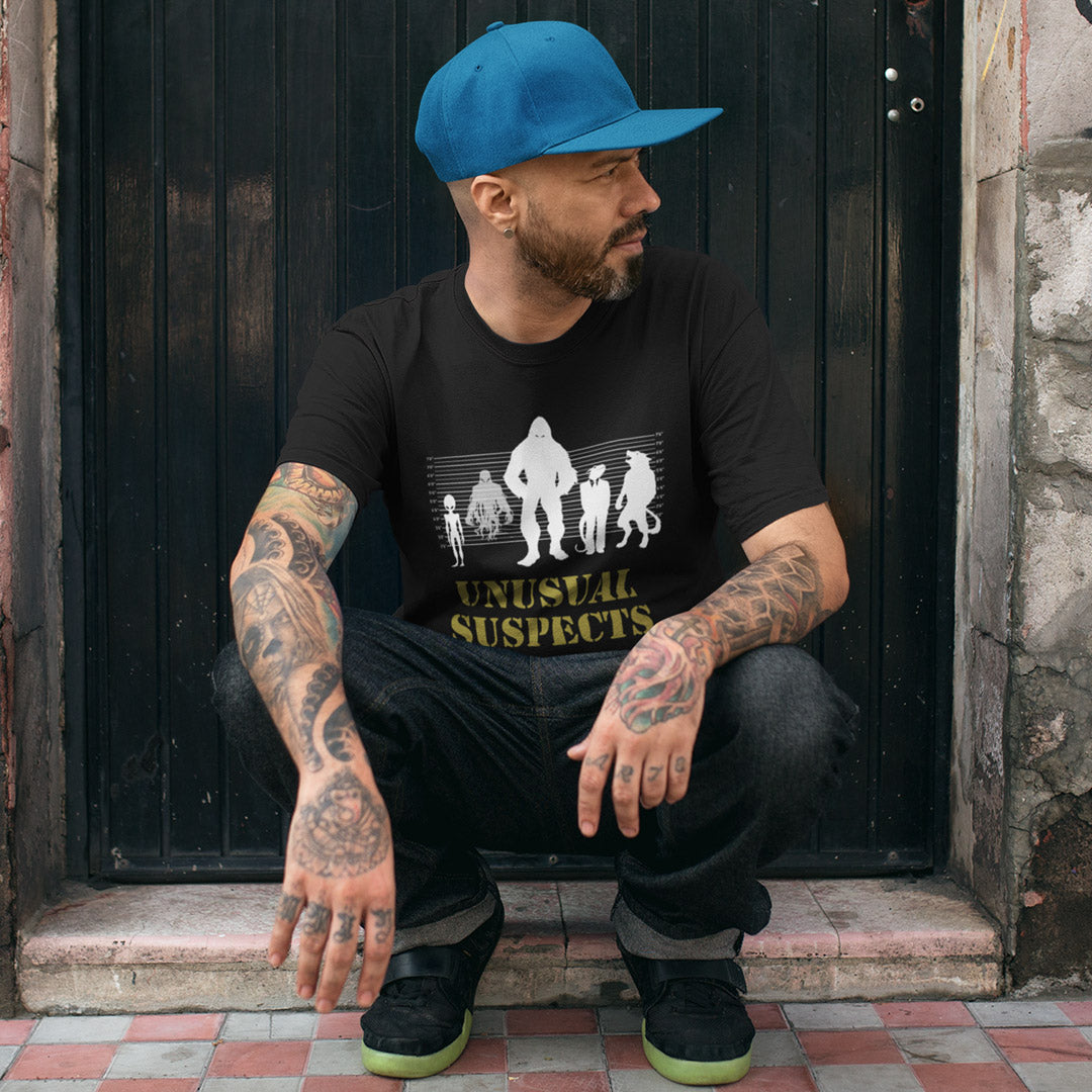 Man in Blue cap, crouched in a doorway, wearing a black t shirt with "UNUSUAL SUSPECTS" text.