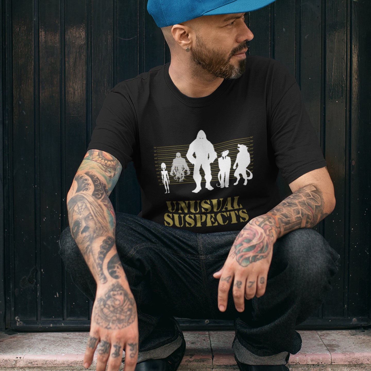 Man wearing a black t-shirt with 'Unusual Suspects' graphic and a blue cap, sitting against a dark wooden wall. A design for movie loving conspiracy theorists. From bigfoot to aliens, these are the personnel responsible for most cover ups and disputed existence. This imaginative stencil art is truly for people who think outside the box. Made from 100% organic ring-spun cotton, this witty conspiracy theory t-shirt is unisex, high-quality, super comfy, and best of all, eco-friendly.