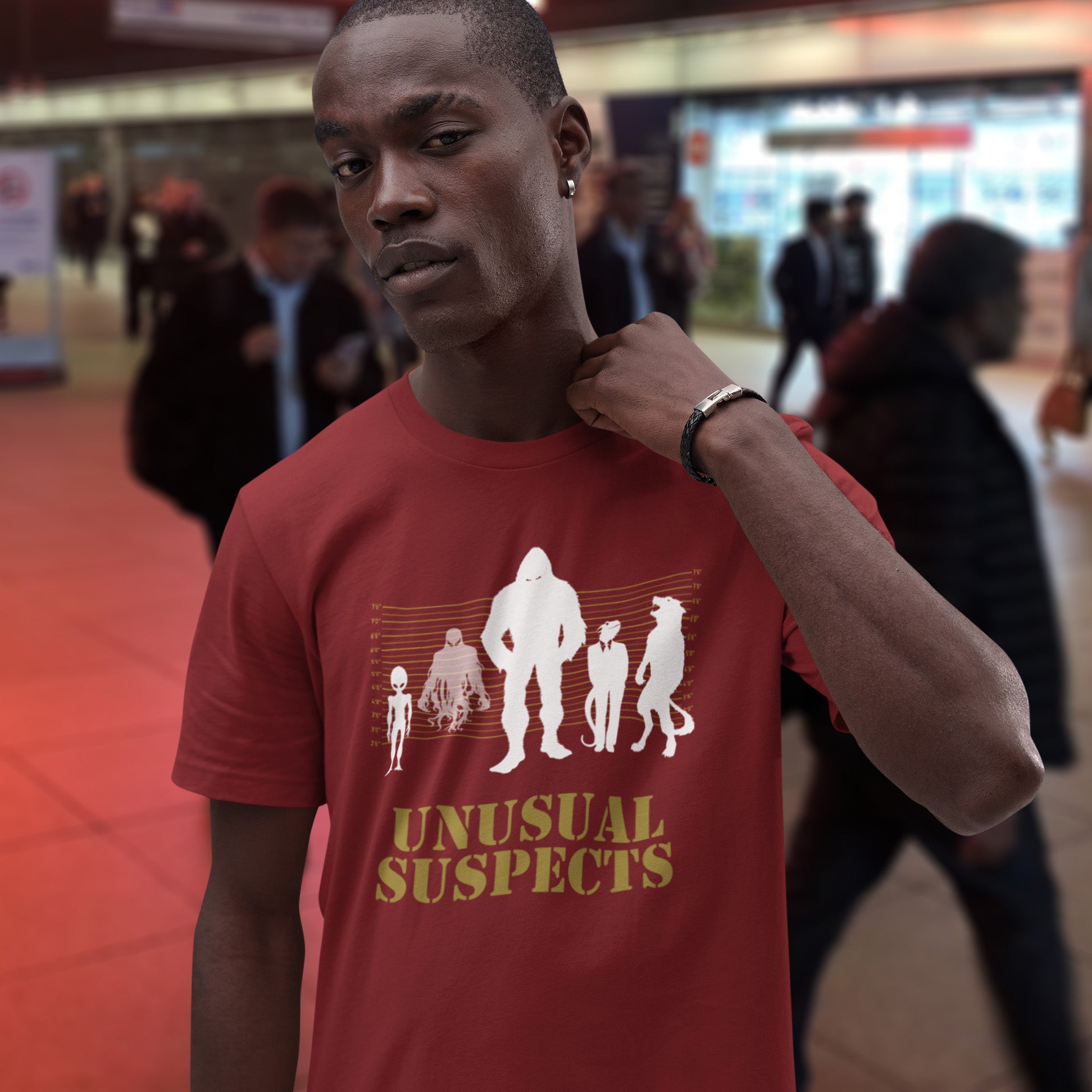 Person wearing a red t-shirt with 'Unusual Suspects' graphic and text in a public indoor setting. A design for movie loving conspiracy theorists. From bigfoot to aliens, these are the personnel responsible for most cover ups and disputed existence. This imaginative stencil art is truly for people who think outside the box. Made from 100% organic ring-spun cotton, this witty conspiracy theory t-shirt is unisex, high-quality, super comfy, and best of all, eco-friendly.