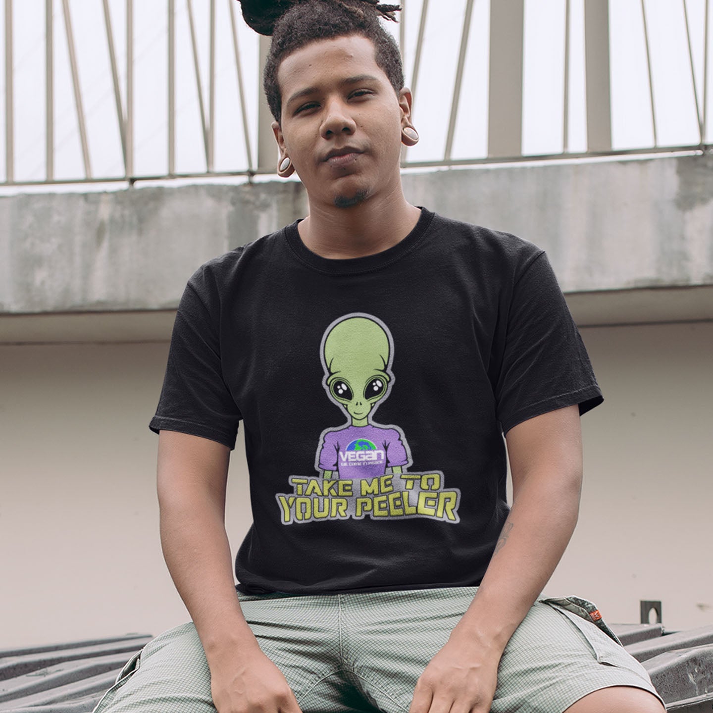 Person wearing a black t-shirt with an alien graphic and text, sitting outdoors. A plant based t-shirt design for anyone that thinks aliens must be vegans, otherwise they would have been farming us years ago. This unique alien design is for all those conspiracy theorists out there. Made from 100% organic ring-spun cotton, this vegan t-shirt is unisex, high-quality, super comfy, and best of all, eco-friendly.