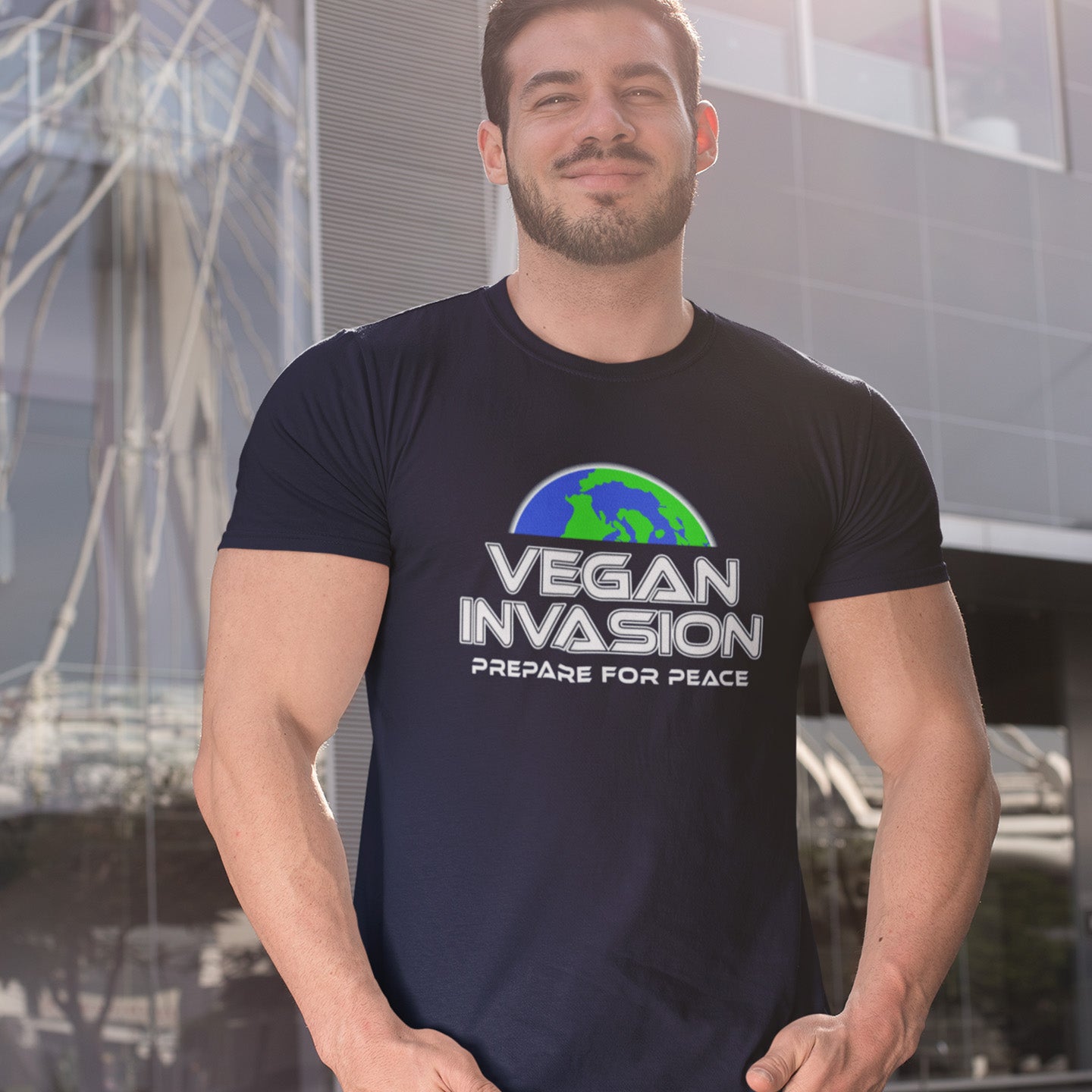 Man wearing a black t-shirt with 'Vegan Invasion' graphic and text, standing in front of a modern building. This vegan meat free t shirt design plays on the idea that other people view vegans as alien. Reassure people that you are a peaceful creature and remind them that you help to save the planet. Made from 100% organic ring-spun cotton, this plant based meat free t shirt is unisex, high-quality, super comfy, and best of all, eco-friendly.