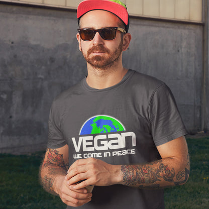 Man wearing an anthracite t-shirt with 'VEGAN WE COME IN PEACE' text and a red cap, standing against a concrete wall. This vegan t-shirt design is for any vegan who feels like they are seen as an alien by others. Reassure people that you are a peaceful creature and want to save the planet. Made from 100% organic ring-spun cotton, this plant based t-shirt is unisex, high-quality, super comfy, and best of all, eco-friendly.