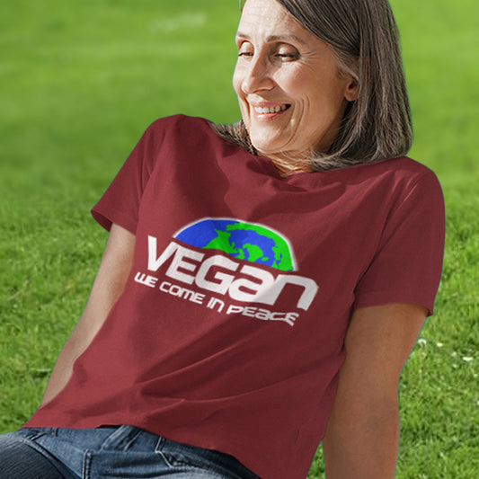 A woman sat on the grass, smiling and wearing a burgundy short sleeved crew neck t-shirt with a graphic of half a globe with the text "VEGAN WE COME IN PEACE" under it.