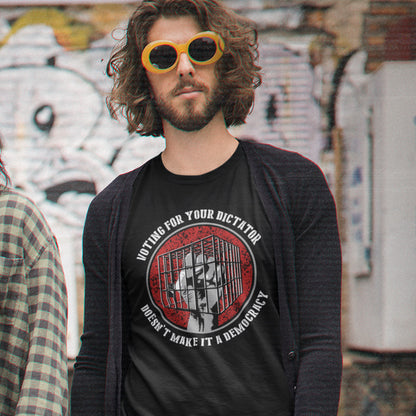 A man with long brown hair and a beard wearing yellow-tinted sunglasses and a black graphic t-shirt under a dark grey cardigan, standing in front of a graffiti-covered wall.