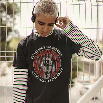 A person with short silver hair wearing a black graphic t-shirt over a black and white striped long-sleeve shirt, with headphones on, standing in front of a white metal fence.