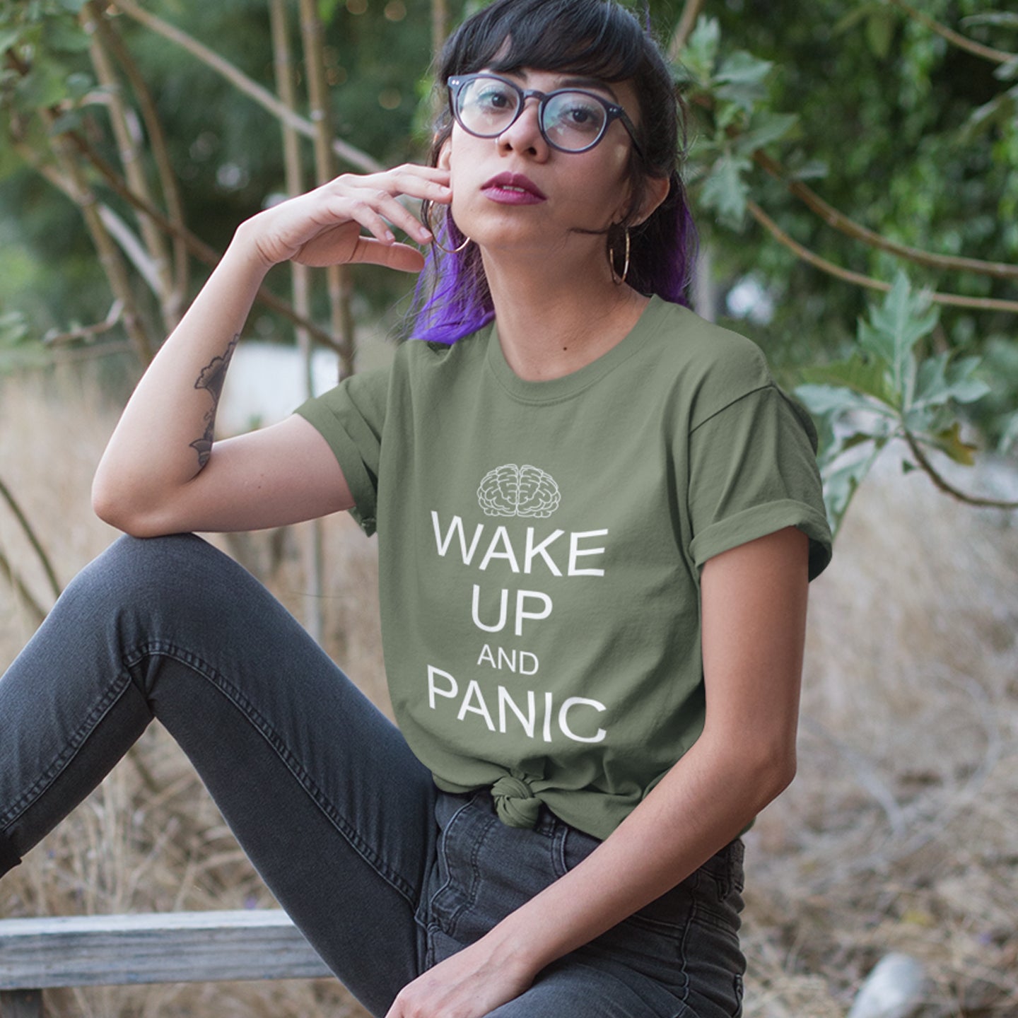 Person wearing a khaki t-shirt with 'Wake Up and Panic' text, sitting outdoors. This satirical 'stay calm' anti-establishment t-shirt suggests we have been calm too long and need of waking up and panic. Keeping calm got us into this mess, it's time to wake up. 100% organic ring-spun cotton, this anti-government t-shirt is unisex, high-quality, super comfy, and best of all, eco-friendly.