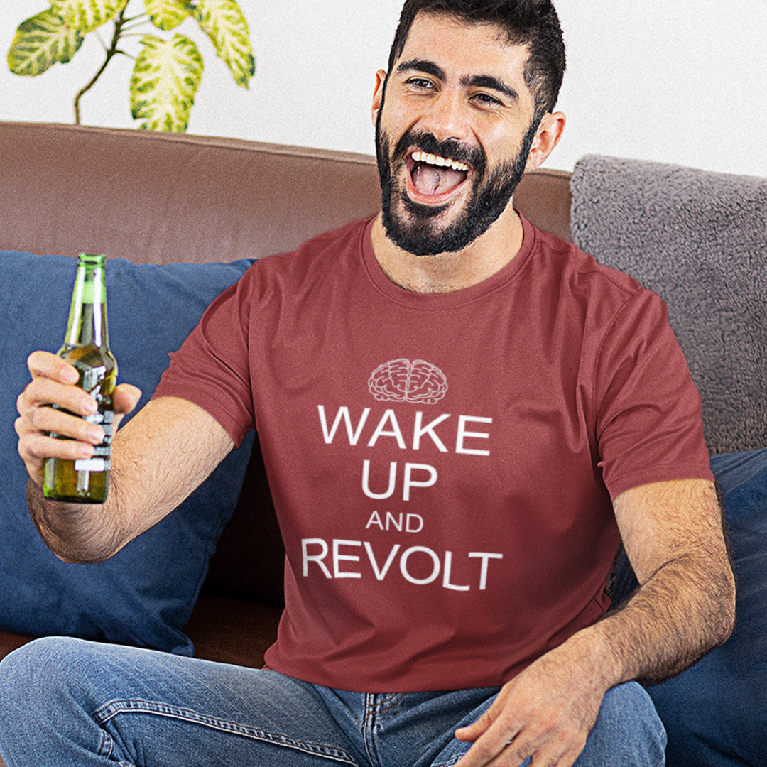 A man sitting on a couch wearing a burgundy t-shirt short-sleeve crewneck t-shirt with a white graphic print on the front. The print features a simple line-art brain icon above the text 'WAKE UP AND REVOLT' in a bold, sans-serif font, styled like the classic 'Keep Calm' poster.