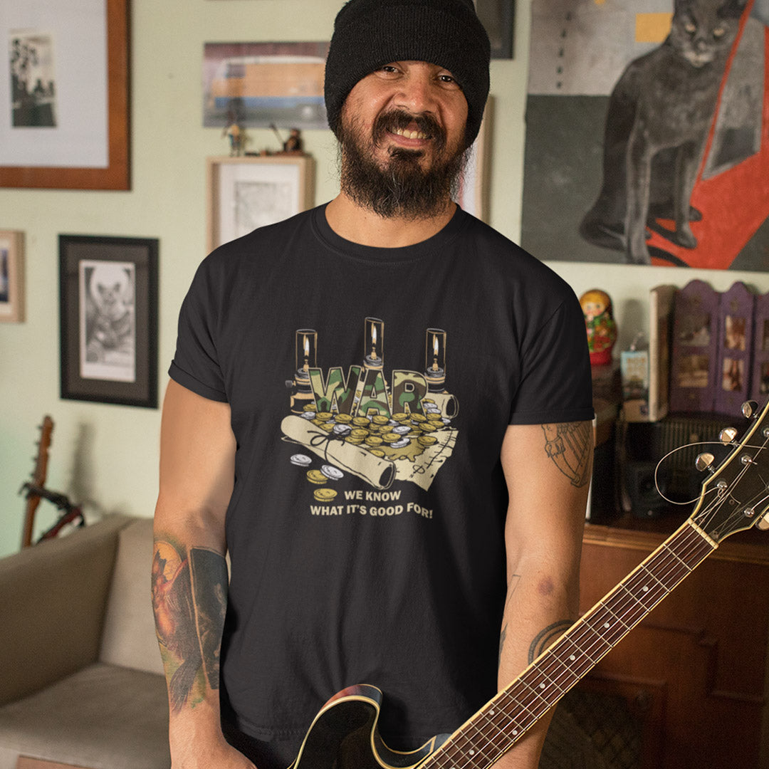 A man wearing a black graphic t-shirt and a black beanie, holding an acoustic guitar in a cozy, decorated room with framed pictures and a cat poster on the wall.