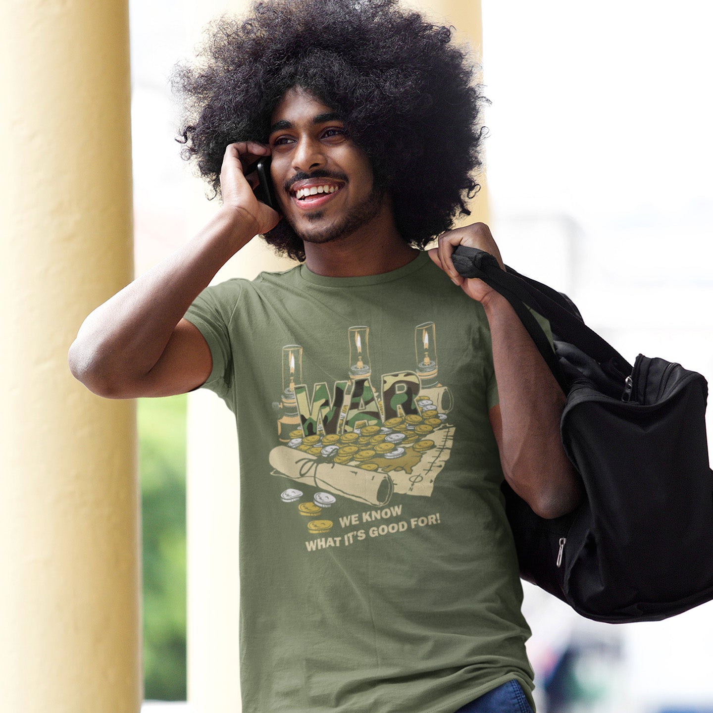 Man wearing a khaki t-shirt with a graphic design, holding a black bag. A satirical anti-war t-shirt suggesting we know what it's good for, money and land deeds for starters. Expose the big business of War with this unique design made for any anti-war demonstration. Made from 100% organic ring-spun cotton, this non conformist t shirt is unisex, high-quality, super comfy, and best of all, eco-friendly.