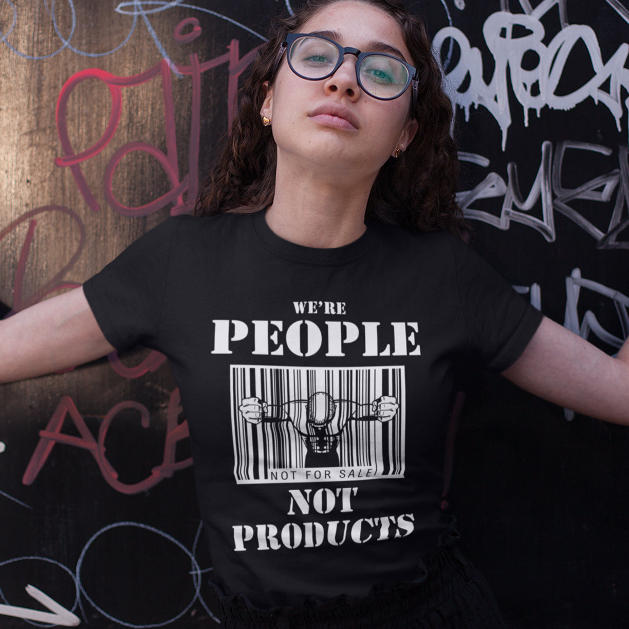 Woman standing against a graffiti wall, wearing a 'we're people not products' t shirt. An anti capitalism t shirt highlighting the dehumanisation of people in a money obsessed society. A fantastic choice for any anti capitalism protest or to just create social awareness. Made from 100% organic ring-spun cotton, this anti consumerism t shit is unisex, high-quality, super comfy, and best of all, eco-friendly.